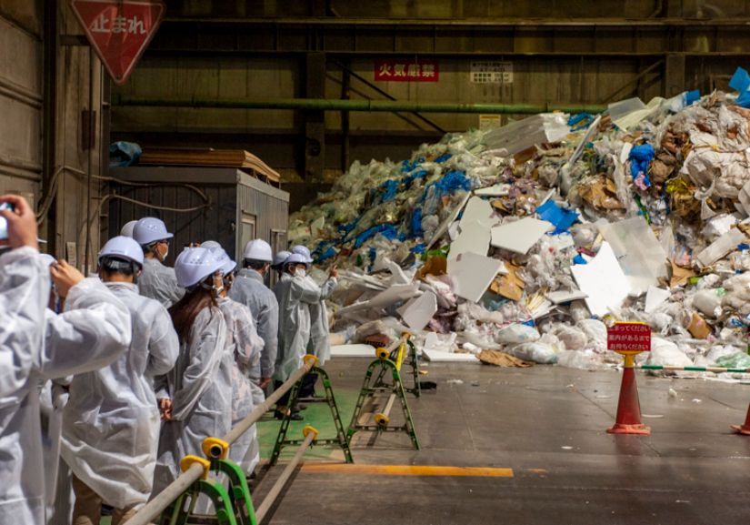 People in white helmets and coats touring a three-meter-high pile of white polystyrene foam and blue plastic rubbish in a warehouse-like building.