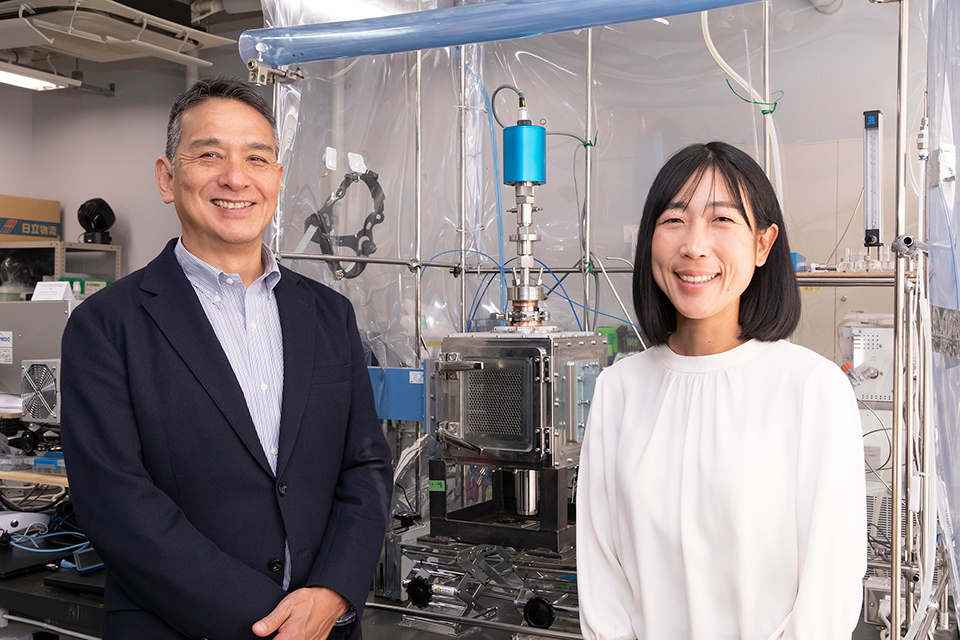 Two people standing in a laboratory setting with various equipment in the background.