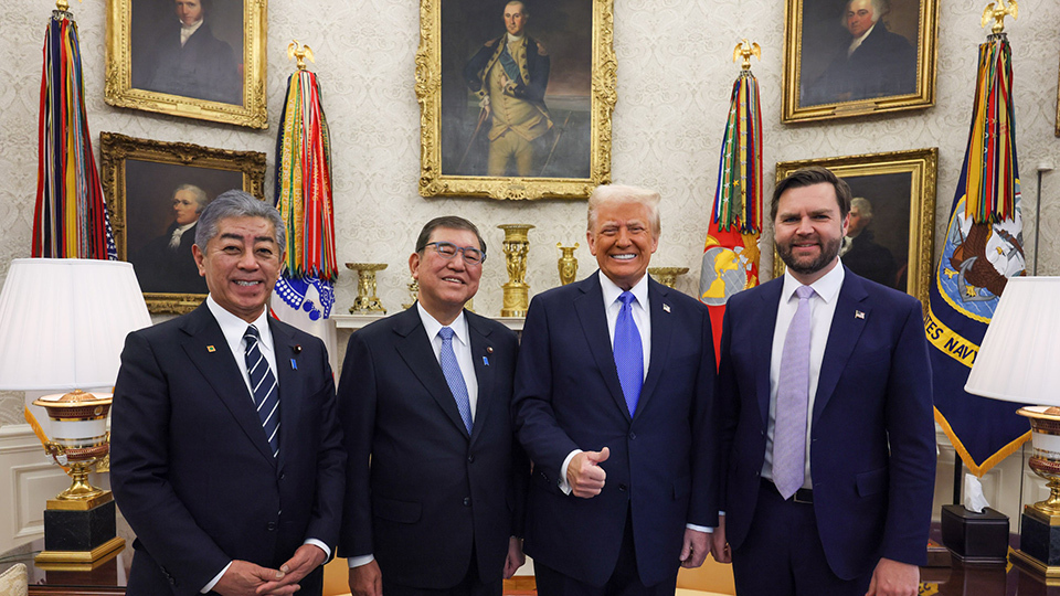Four men in suits stand in a room adorned with flags and portraits in gilded frames.