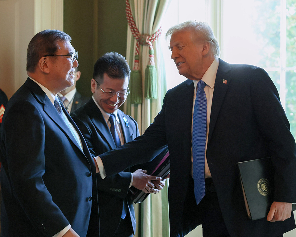 Two men in suits engage in conversation, with one rearching out towards the other,  third man smiles while holding folders behind them.