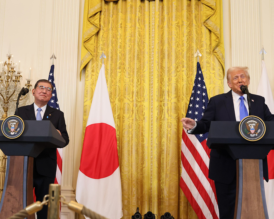 Two men standing at podiums during a press conference, with the United States and Japanese flags prominently displayed behind them.