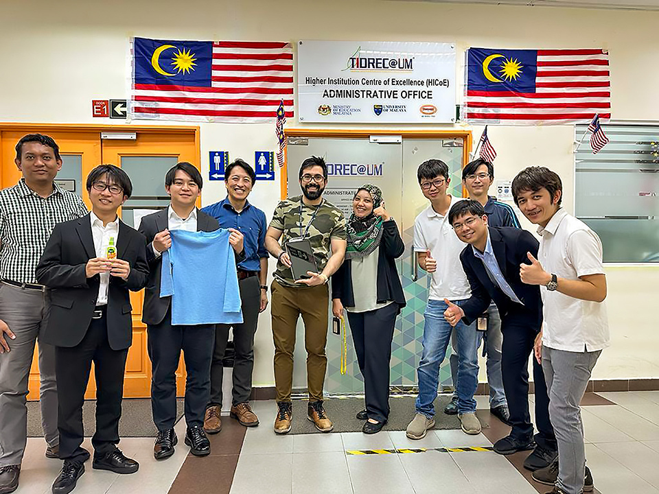 A diverse group of individuals.stands in front of a room displaying two Malaysian flags, at the Tropical Infectious Diseases Research & Education Centre (TIDREC) in Malaysia.
