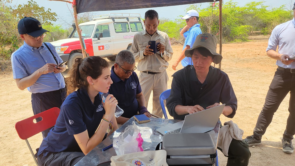 A man shows his laptop to an NGO staff member, with others around the table, in an outdoor setting.