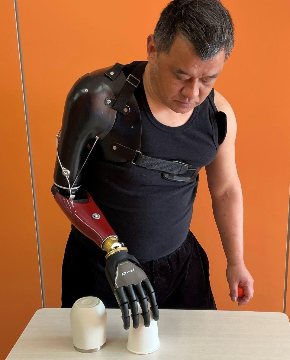 A man wearing a black sleeveless shirt using a bionic prosthetic arm to manipulate two small cups.