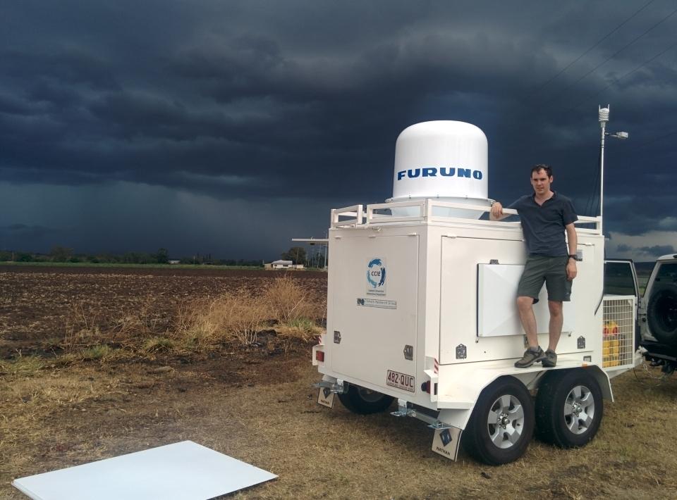 A compact weather radar mounted on a mobile observation vehicle,with a man posing in front and dark clouds in the background.