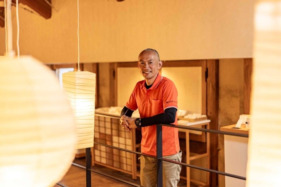 Man in a orange shirt posing on a railing, with hanging lamps made from washi paper hanging in the foreground