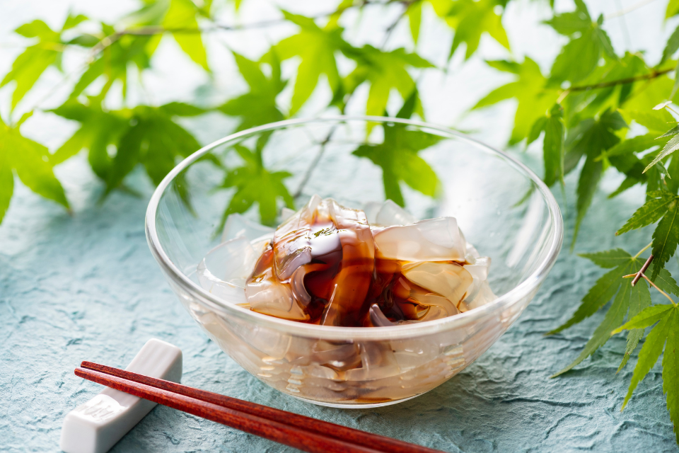 A glass bowl filled with clear jelly-like noodles, covered in a dark sweet sauce and framed by green maple leaves