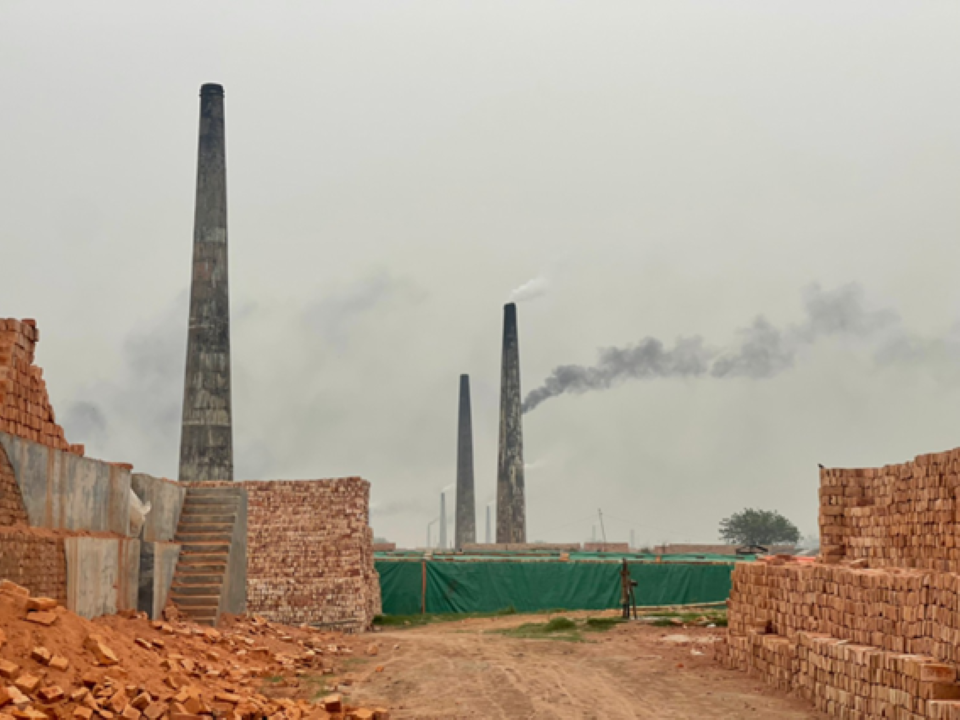 A fired brick factory, with grey smoking chimneys and piles and stacks of bricks in the foreground