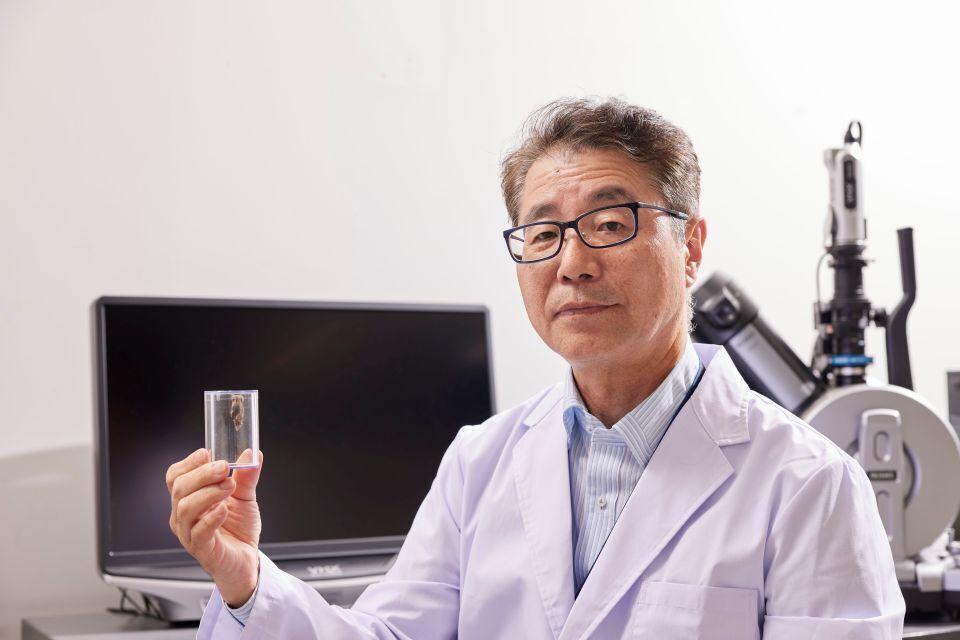 An older Japanese man in a white lab coat, holding a container with a bagworm inside
