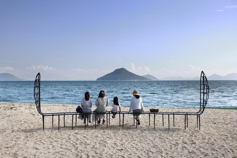 Three women and one child seated on a bench-like artwork, Place for Sea Dreamers, on a sandy beach overlooking the ocean and a island in the distance. 