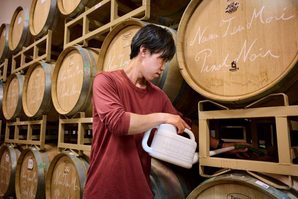 A young man topping up large wooden wine barrels