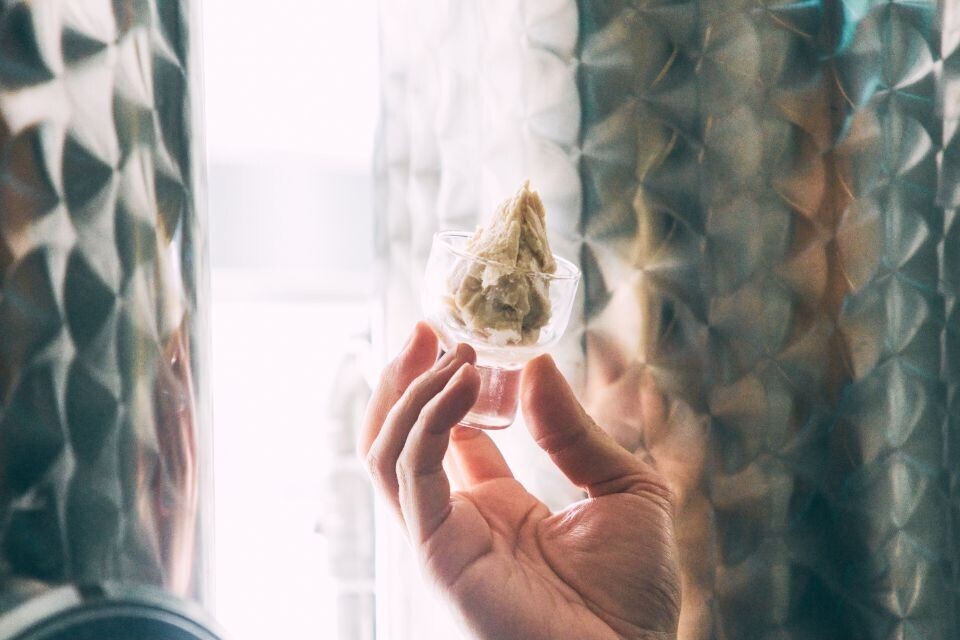 A small glass filled with white sake lees, being held up in front of metal tanks