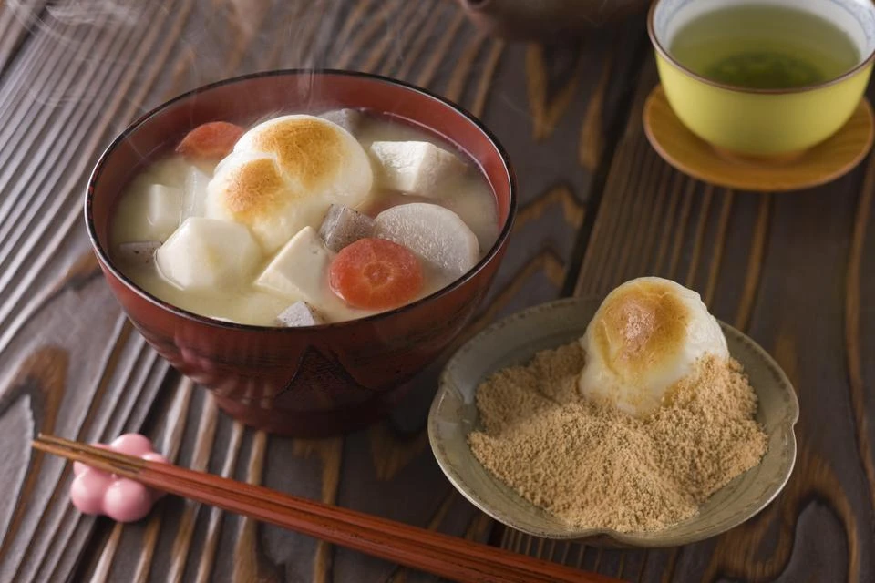 Image of zoni, a soup willed with mochi rice cakes, with a rice cake on the side being dipped in roasted soybean powder